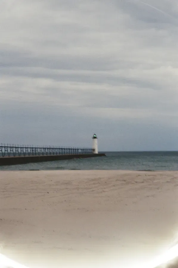 Beach scene with Manistee North Pierhead lighthouse at the end of a pier under a cloudy sky.