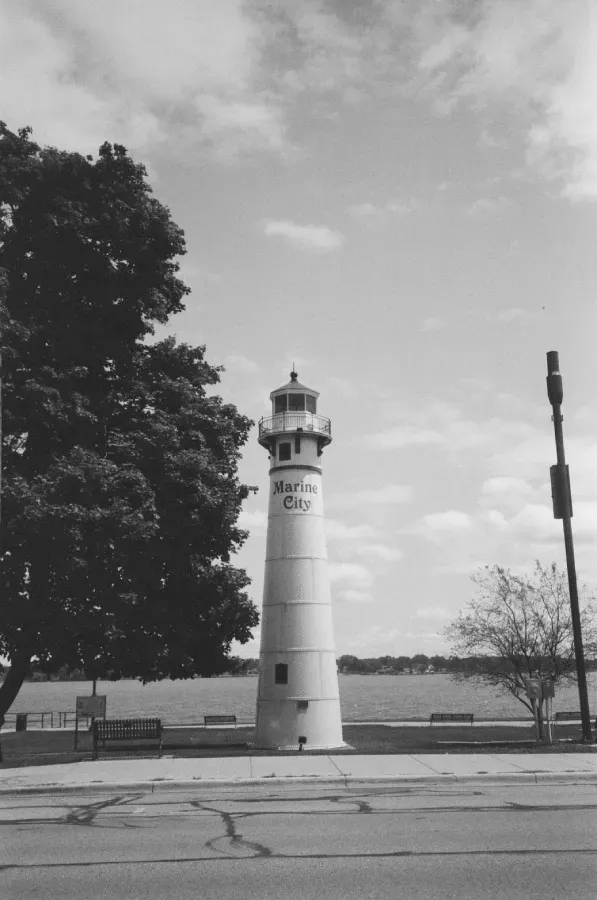 Black and white film photo of Marine City lighthouse. Tree on the left, sky with clouds overhead.