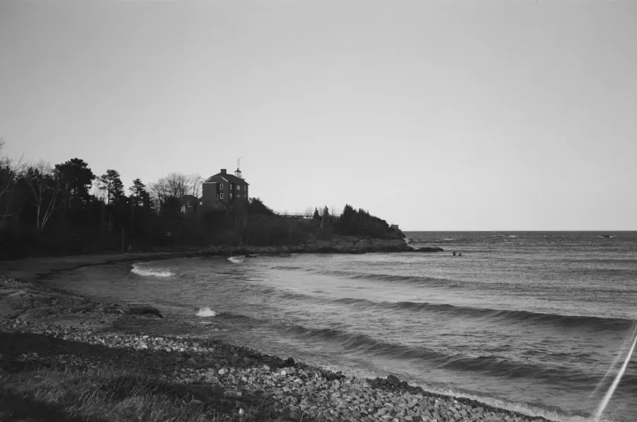 Black and white photograph of a beach with waves. Trees and a building are on the right.