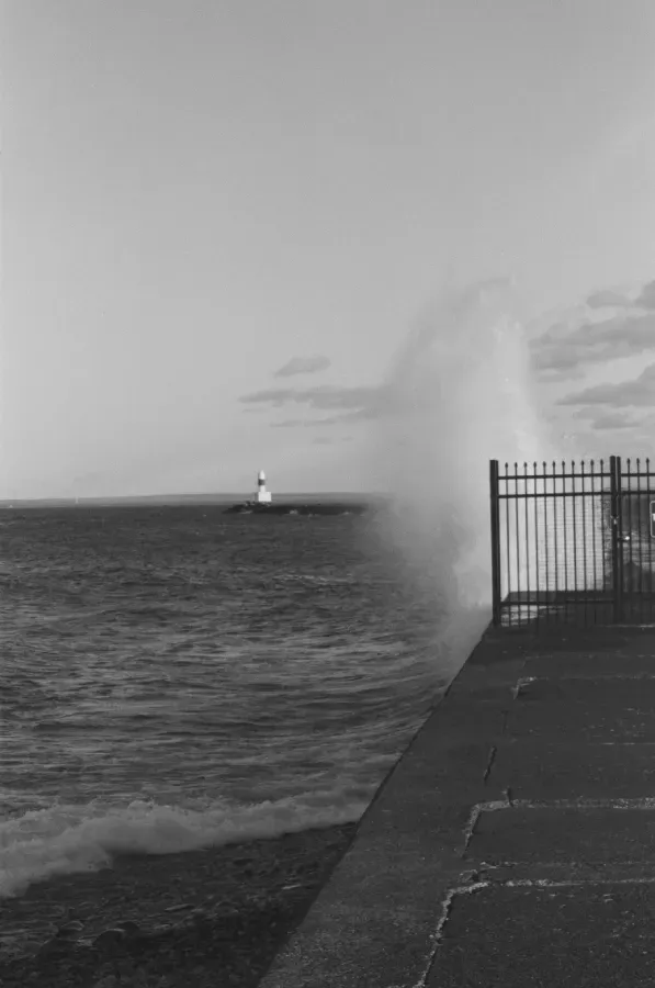 Black and white shot of Marquette Breakwater lighthouse in the distance, waves crashing against a seawall.