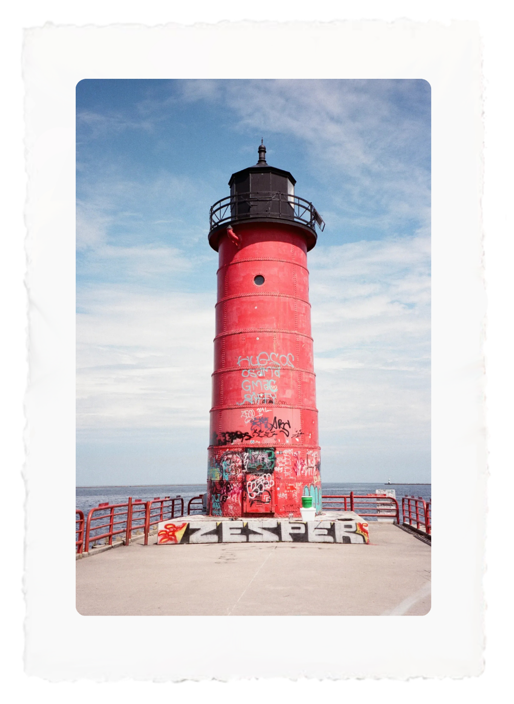 Red lighthouse on a pier under a blue sky, with red railings and graffiti at the base