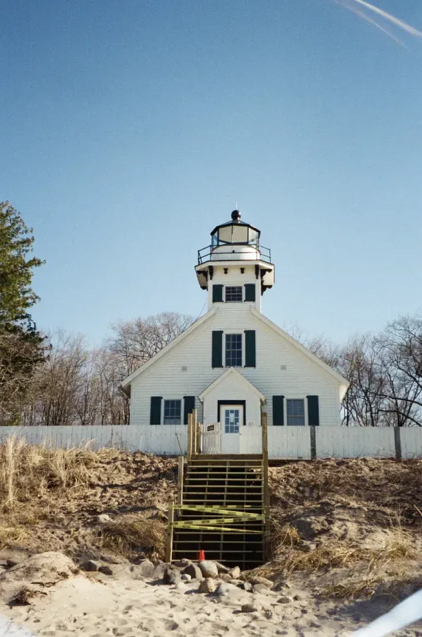 White lighthouse with black lantern room, against a blue sky, accessed by wooden stairs on a sandy bank.
