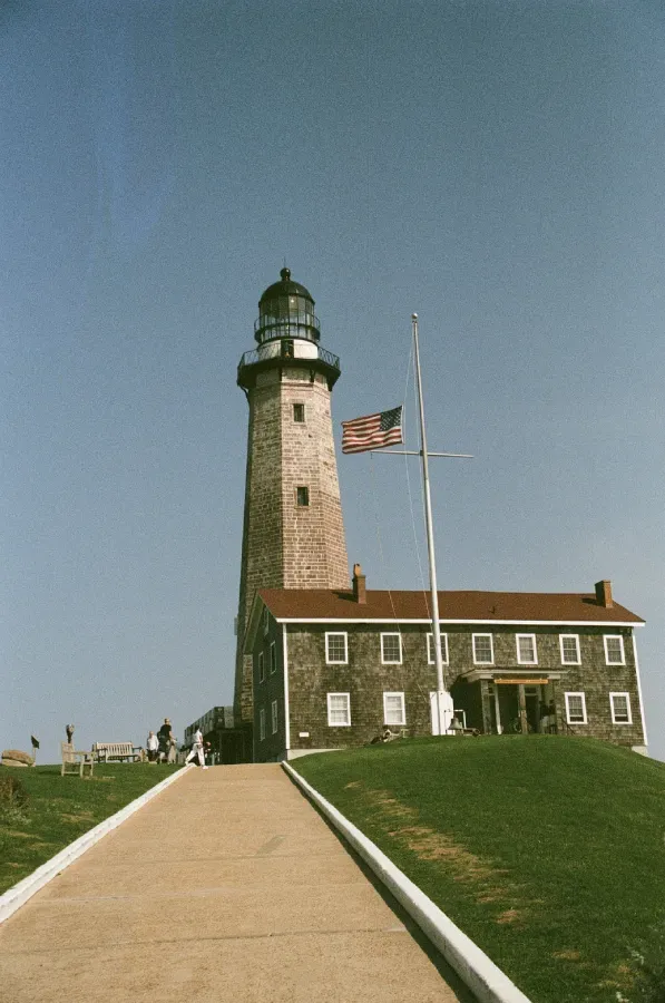 Montauk Point Lighthouse on a hill with an American flag. Brick building and paved path leading up. Blue sky.