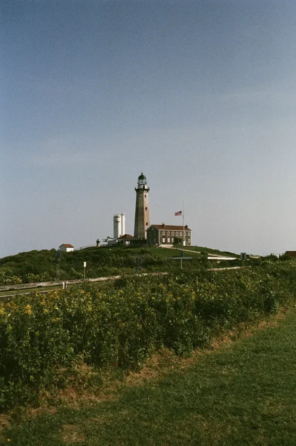 Montauke Point Lighthouse on a hilltop, with a clear blue sky. Green shrubs and grass in the foreground.