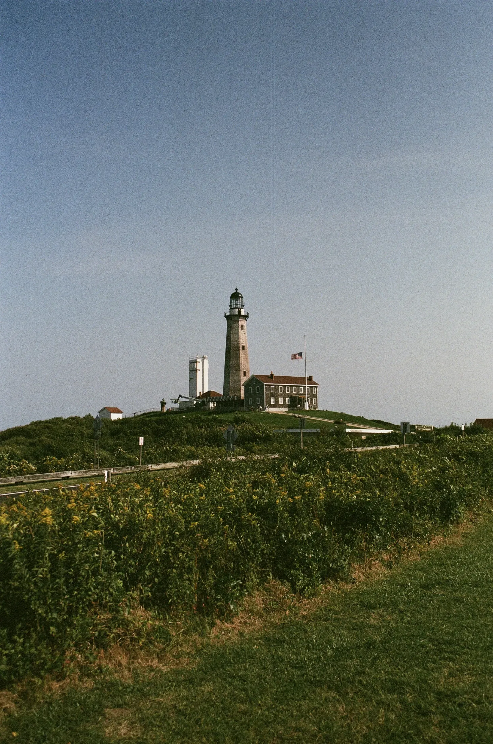Montauke Point Lighthouse on a hilltop, with a clear blue sky. Green shrubs and grass in the foreground.