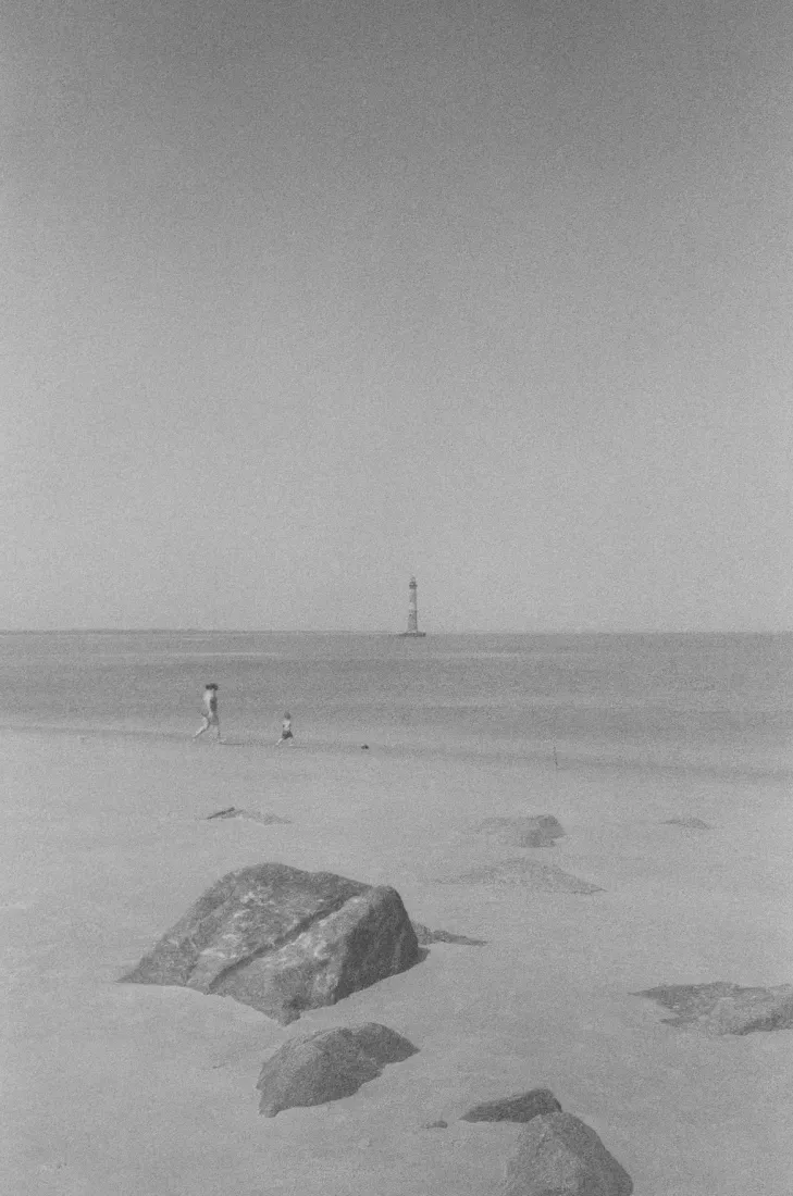 Grayscale beach scene with a lone tent in the sand and a distant lighthouse on the horizon