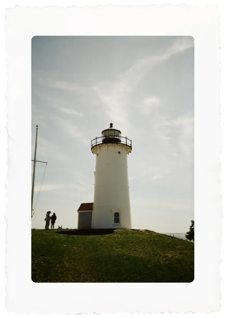 White lighthouse on a grassy hill under a cloudy sky, with two small people standing nearby