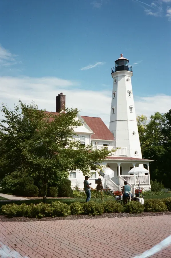 A white lighthouse stands at the end of a stone jetty extending into the ocean under a pale, sunny sky.
