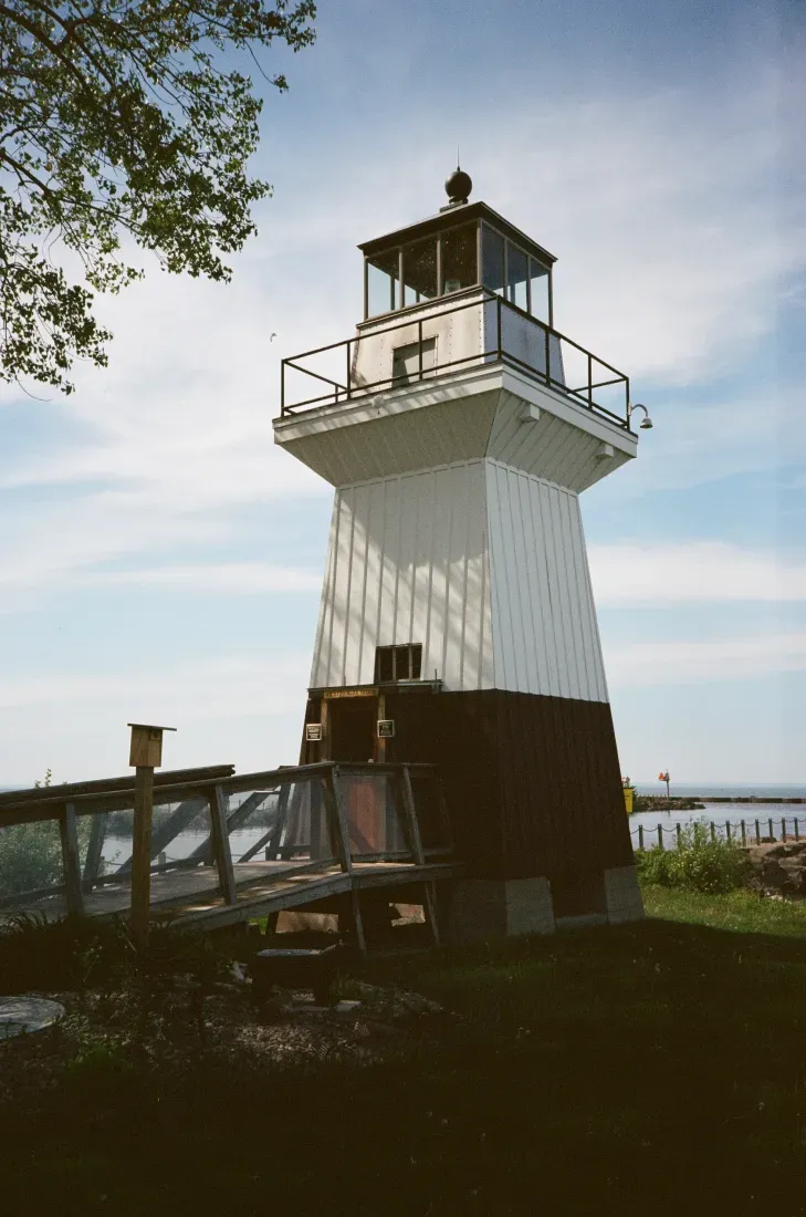 Oak Orchard lighthouse with a dark base and glass-top lantern, standing near water under a blue sky.