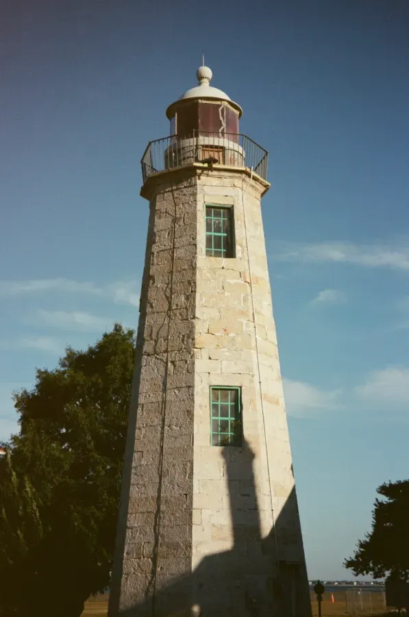 The Old Point Comfort Stone lighthouse under a clear blue sky, with a tree to the left casting a shadow.