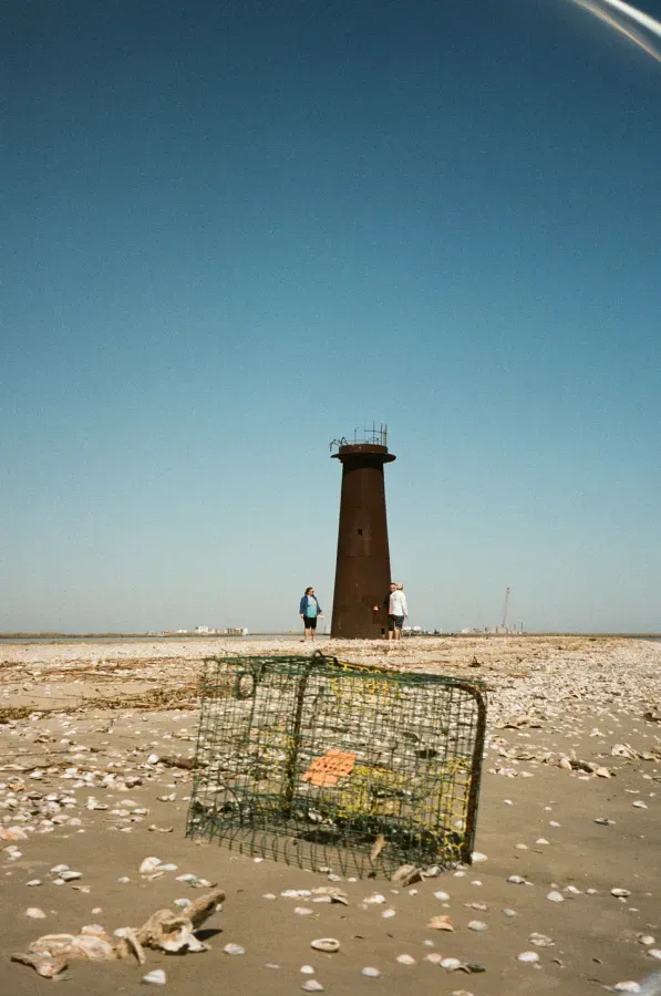 Rusty lighthouse on a sandy shore; two people stand near it, crab trap in foreground, blue sky.