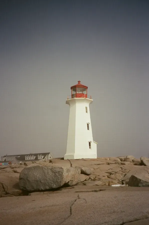 Peggy's Cove lighthouse with a red lantern room stands on a rocky coastline against a hazy, overcast sky