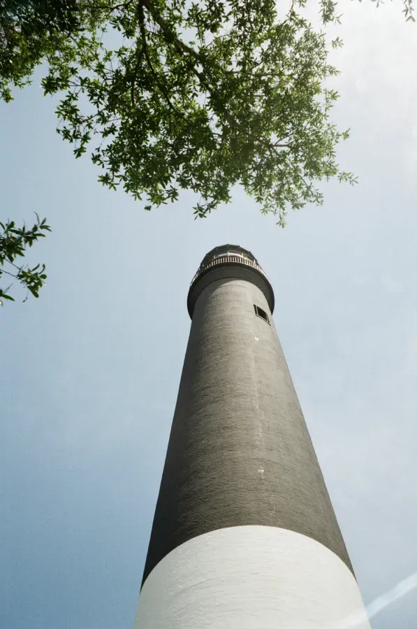 The Pensacola lighthouse, viewed from below, against a blue sky, with green tree branches in the upper frame.