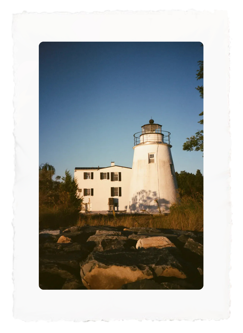 White lighthouse beside a small building under a blue sky, framed by trees and rocks in warm light
