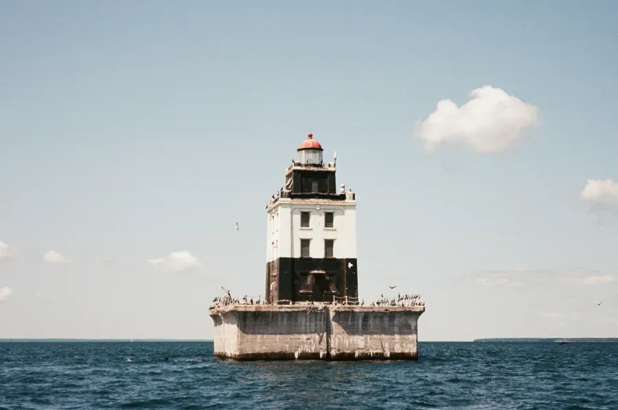Poe Reef Lighthouse on a square foundation in the ocean, blue water and sky, with a small cloud.