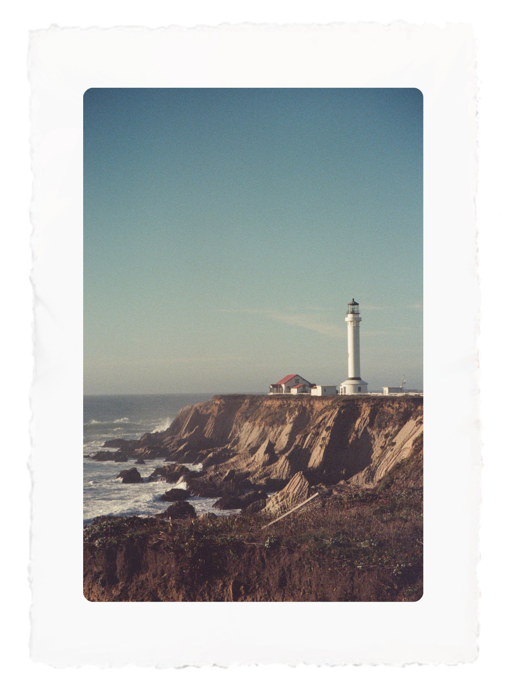 Lighthouse on a rocky seaside cliff under a clear blue sky, with ocean waves below.