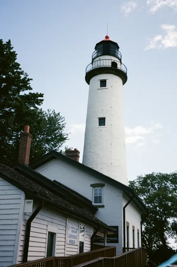 Point Aux lighthouse tower with attached white building and black roof. Green trees and blue sky.
