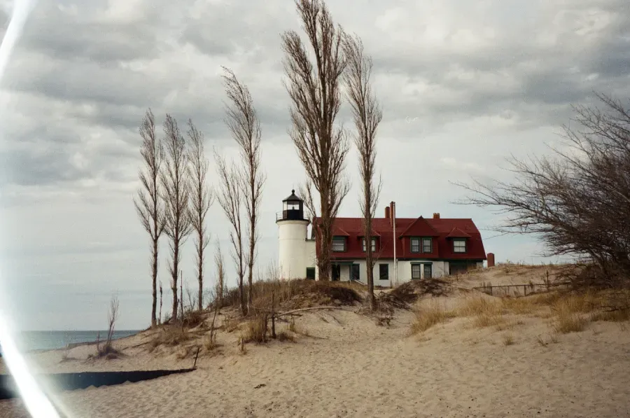Point Betsie Lighthouse with red roof and white tower on sandy dune with bare trees under cloudy sky.