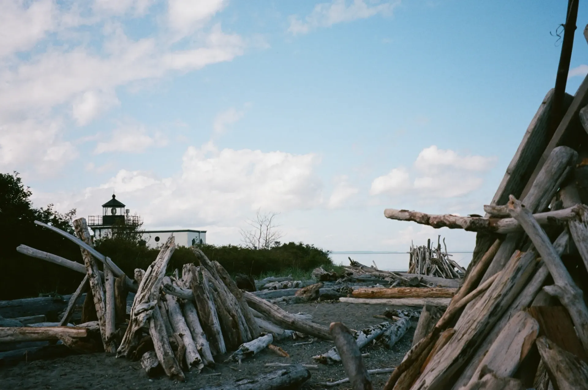 Driftwood structures on a beach with a Point No Point lighthouse in the distance under a blue sky.