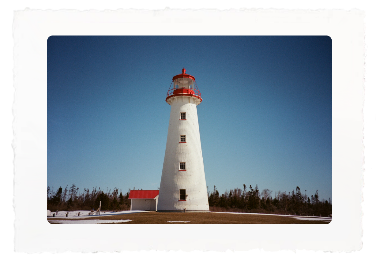 White lighthouse with a red top against a clear blue sky, surrounded by snow and trees