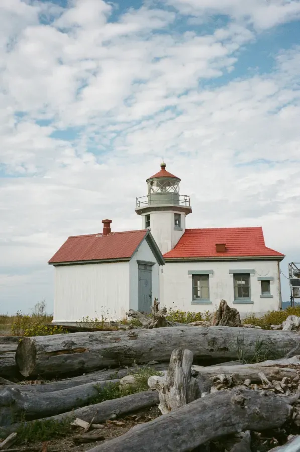 Point Robinson lighthouse with red roof under a cloudy blue sky. Driftwood logs in foreground.