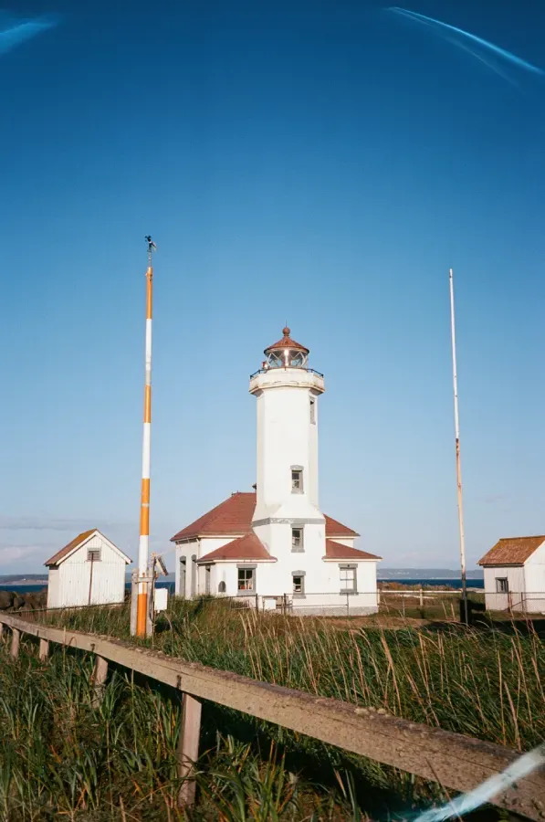 Charlee Remitz smiling wearing sunglasses on their head poses in front of a lighthouse in the ocean under a clear blue sky.