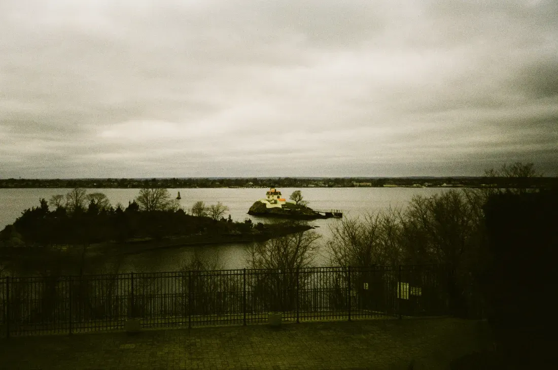 Film photo of Popham Rocks lighthouse in a calm sea under a cloudy sky, viewed through dark foreground shrubs and fence