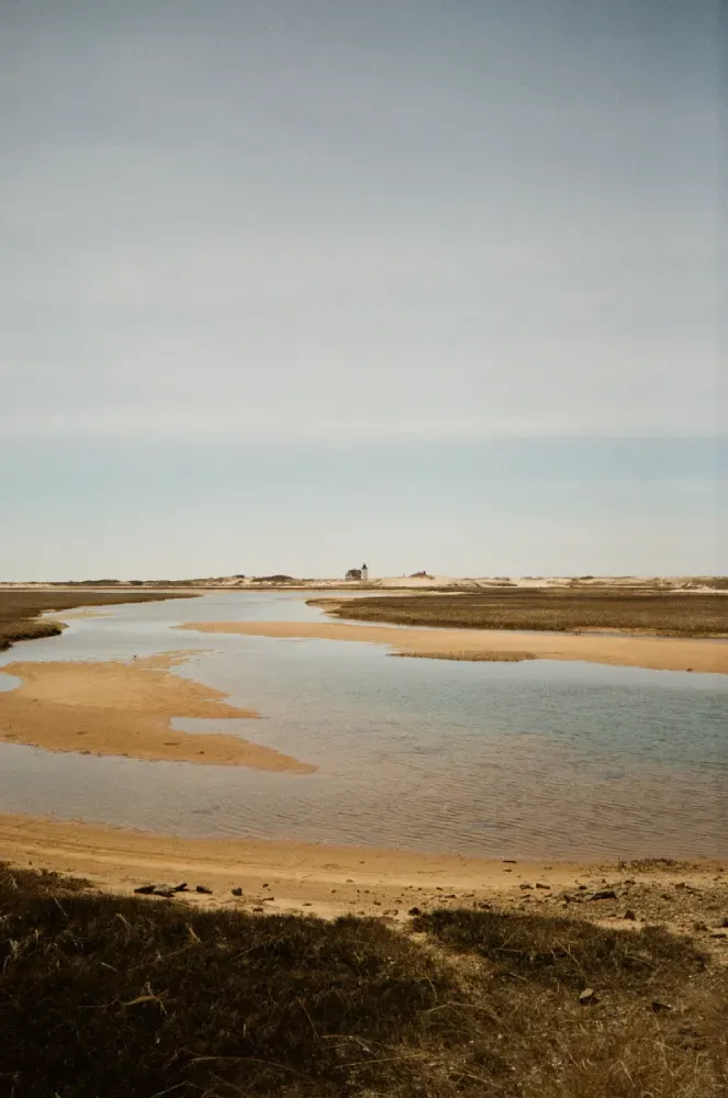 Film photo of Race Point lighthouse in the background of a sandy riverbank with a shallow winding stream under a pale cloudy sky