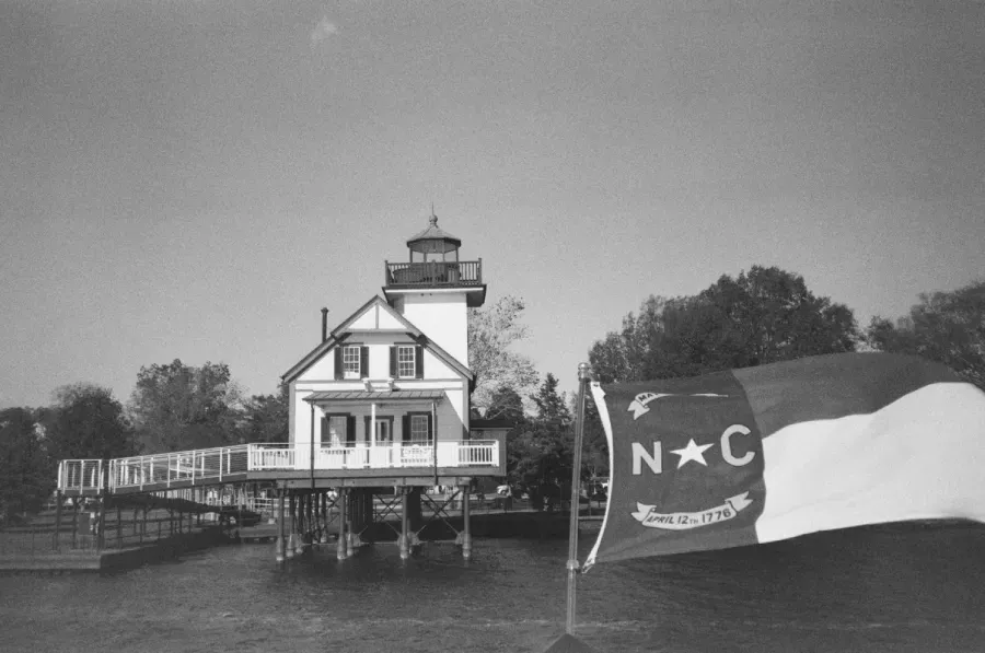Black and white film photograph of Roanoke River lighthouse on stilts over water, with North Carolina flag waving