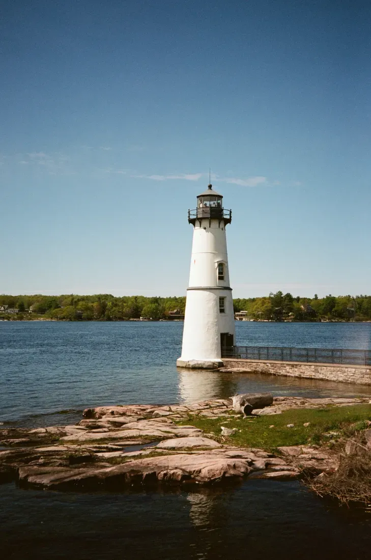 Rock Island lighthouse on a rocky shore beside calm blue water under a clear sky.