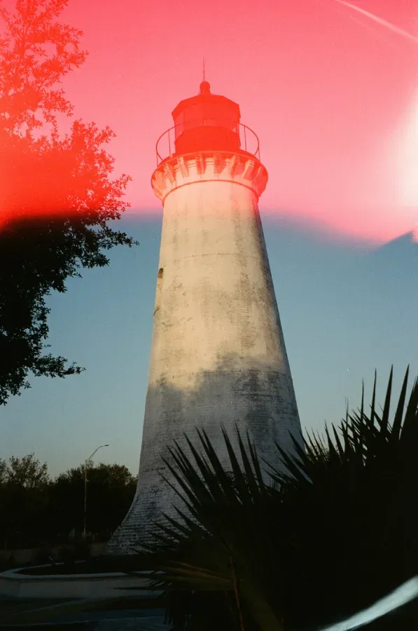 Round Island lighthouse against a blue sky, with red lens flare and dark foliage in foreground.