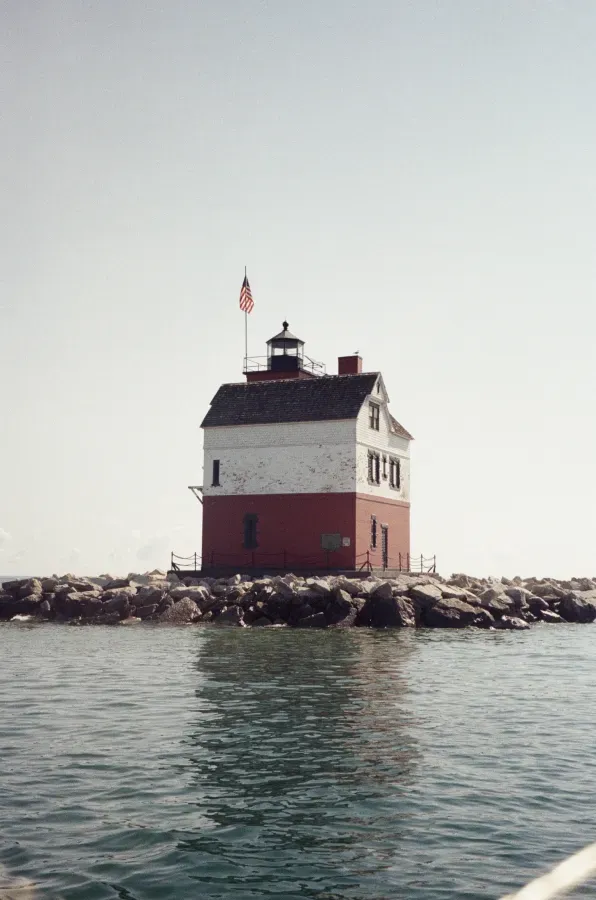 Round Island Michigan Lighthouse on a rocky island, red and white building, American flag on top, water in foreground.