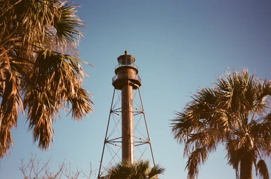 Sanibel Island Lighthouse with openwork metal tower, topped with a light, flanked by palm trees against a blue sky.