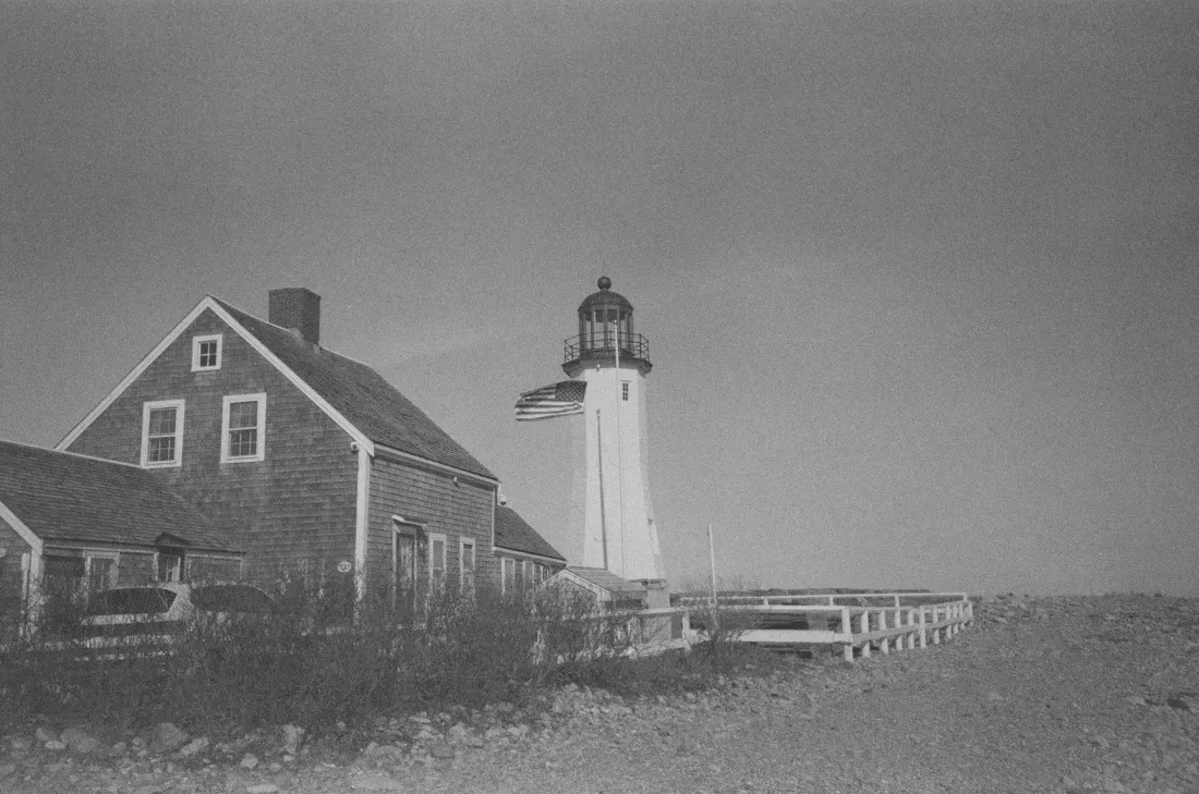 Black-and-white photo of scituate lighthouse beside a weathered house on a rocky coast under a hazy sky