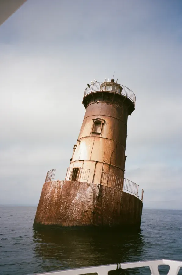 A weathered, rusty brown lighthouse stands alone in the middle of calm, open water under a pale sky.