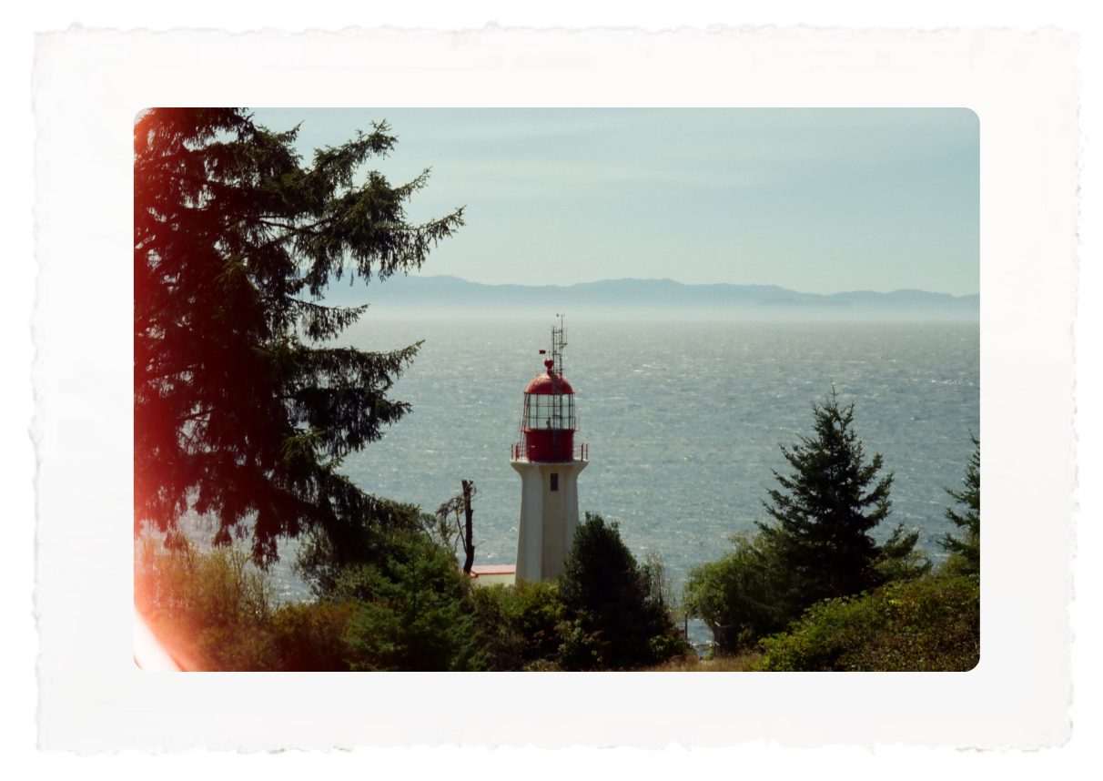 Lighthouse with red top by a calm sea, framed by trees and distant hazy mountains