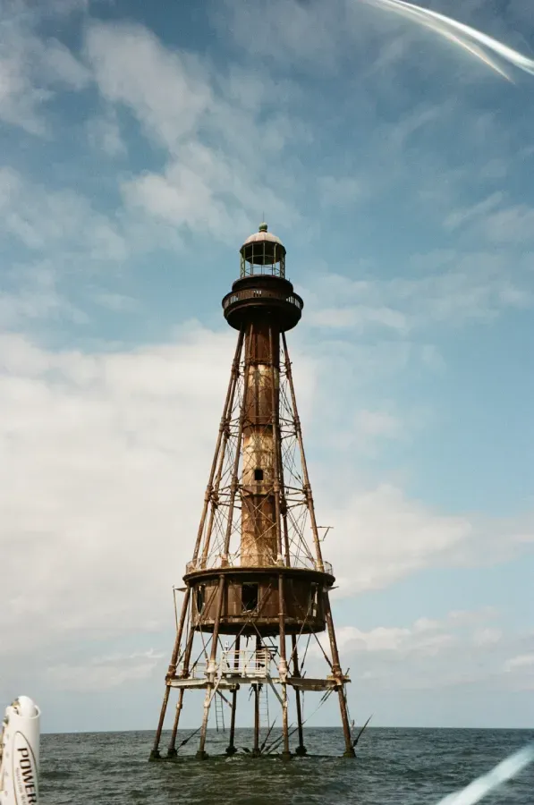 Rusty, open-frame Ship Shoal Lighthouse rising from ocean under a cloudy blue sky.