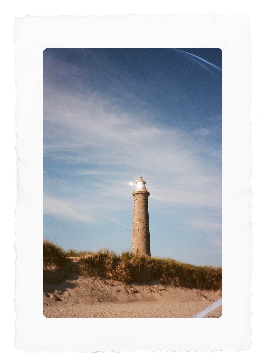 Lighthouse on sandy dunes under a blue sky, framed by a white photo border