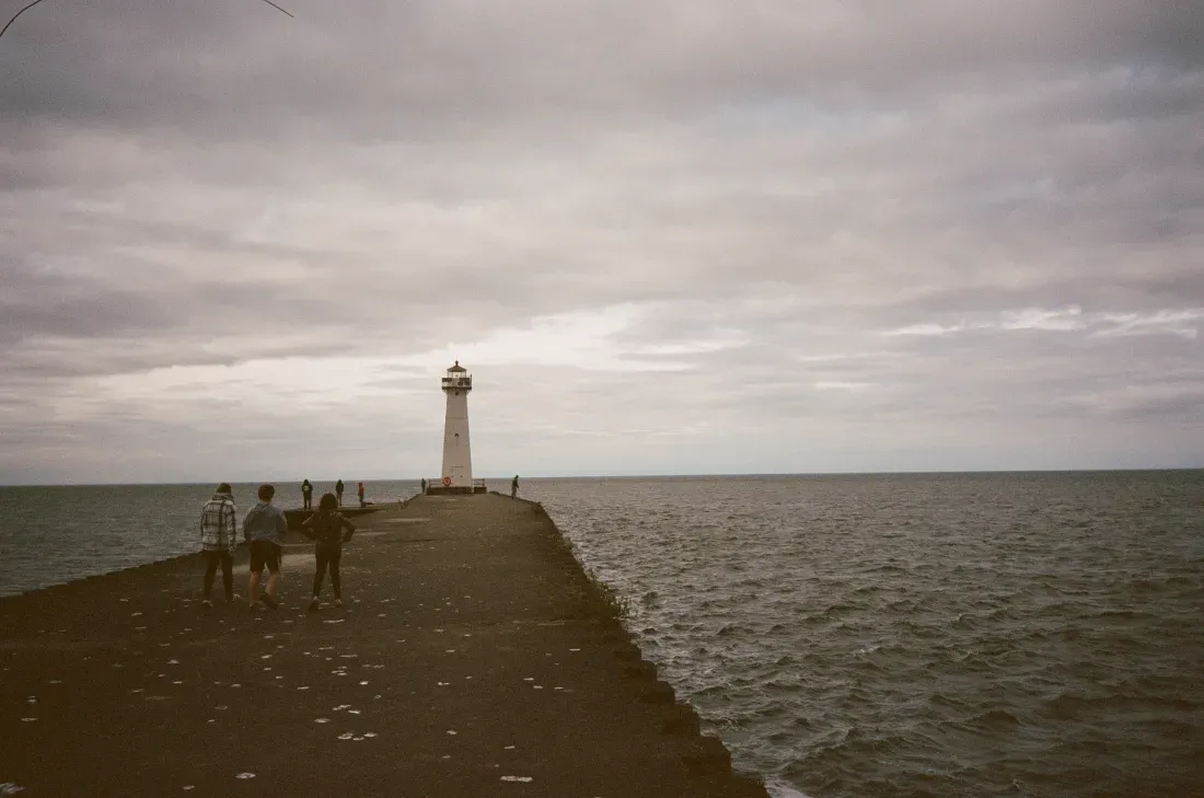 Sodus Bay Lighthouse at the end of a stone pier under a cloudy sky, with a few people walking by the sea.