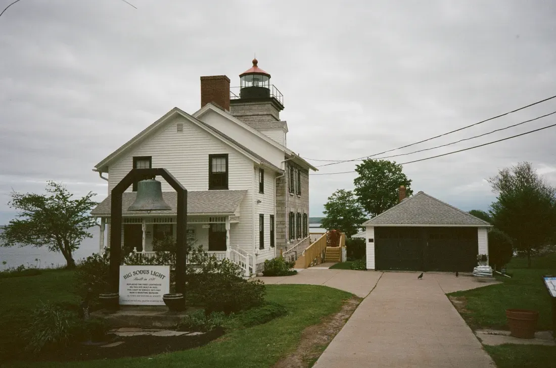 Historic Sodus Bay lighthouse with a small sign and driveway under cloudy skies