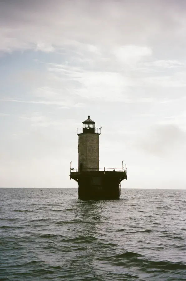 Solomons Lump Lighthouse in the sea, gray stone tower, black base, cloudy sky, calm water.
