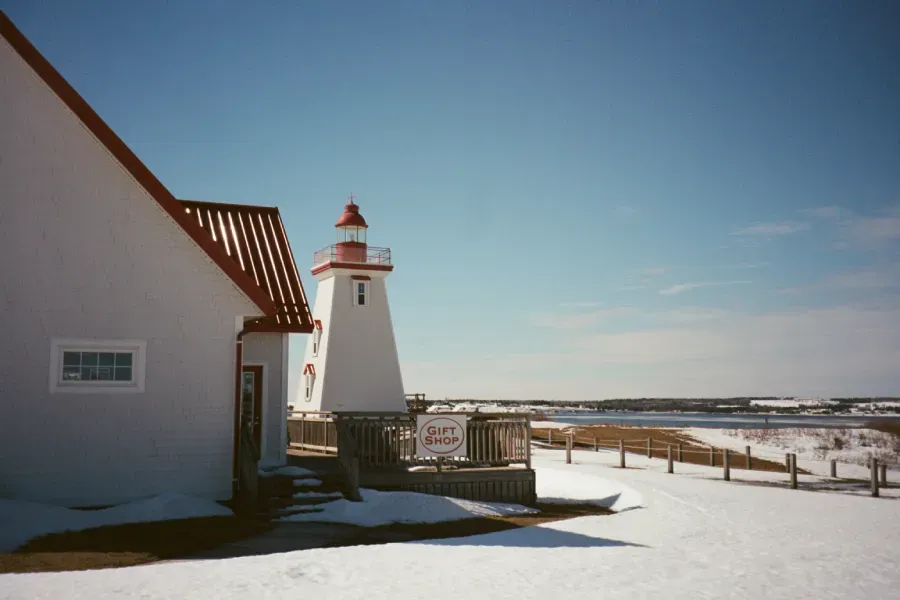 Souris lighthouse with a red roof stands beside a building on a snow-covered shore under a clear blue sky.