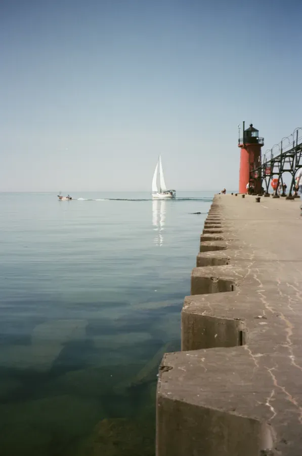 A lighthouse and concrete pier extend into calm water. A sailboat moves away in the distance on a sunny day.