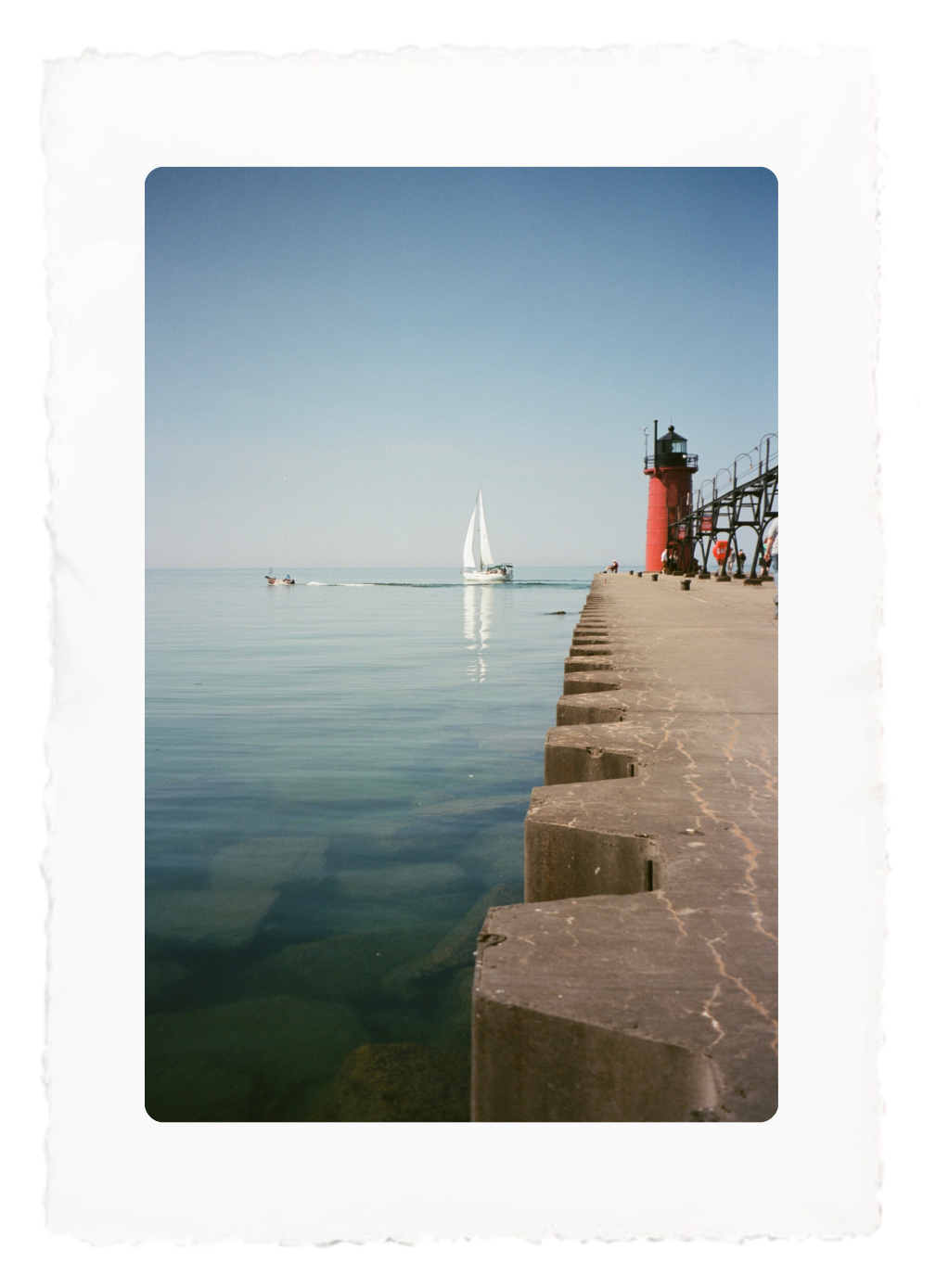 Calm harbor with a white sailboat, stone pier, and red lighthouse under a clear blue sky