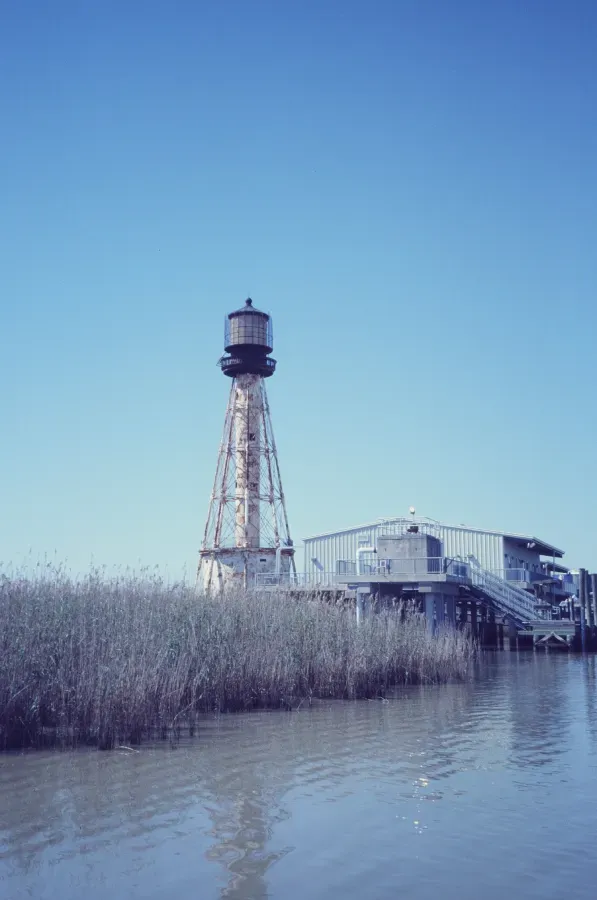 Tall metal lighthouse next to a wooden building on a body of water, reeds in foreground, blue sky.