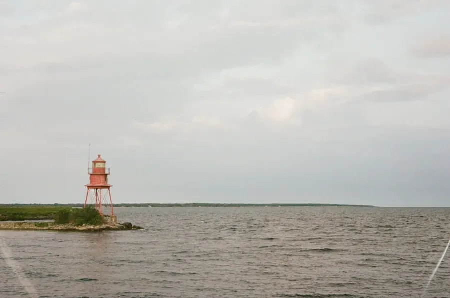 Sputnik Dink stands on a small, grassy point of land extending into a large, calm body of water under a cloudy sky.