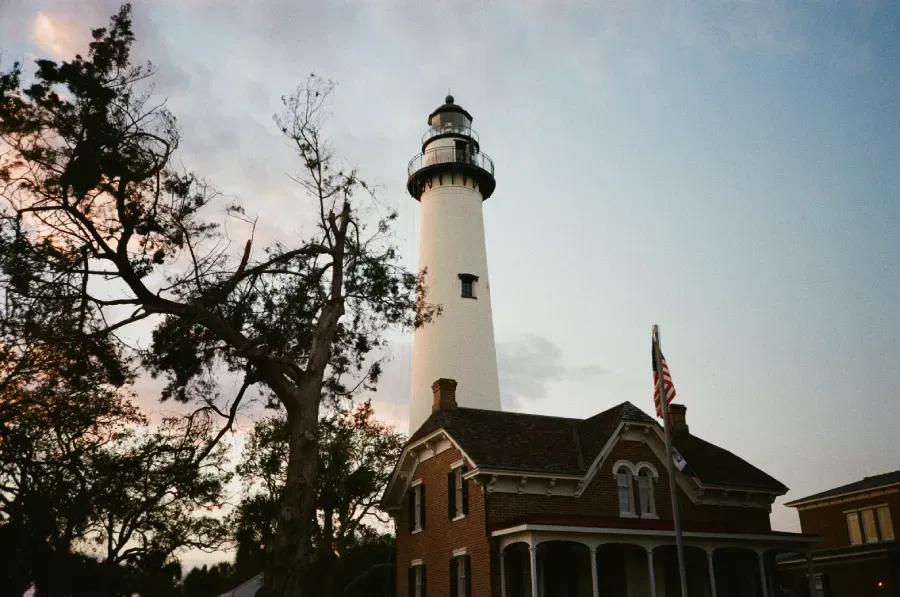 St Simons lighthouse and attached red brick building against a dusky sky, trees on the left.