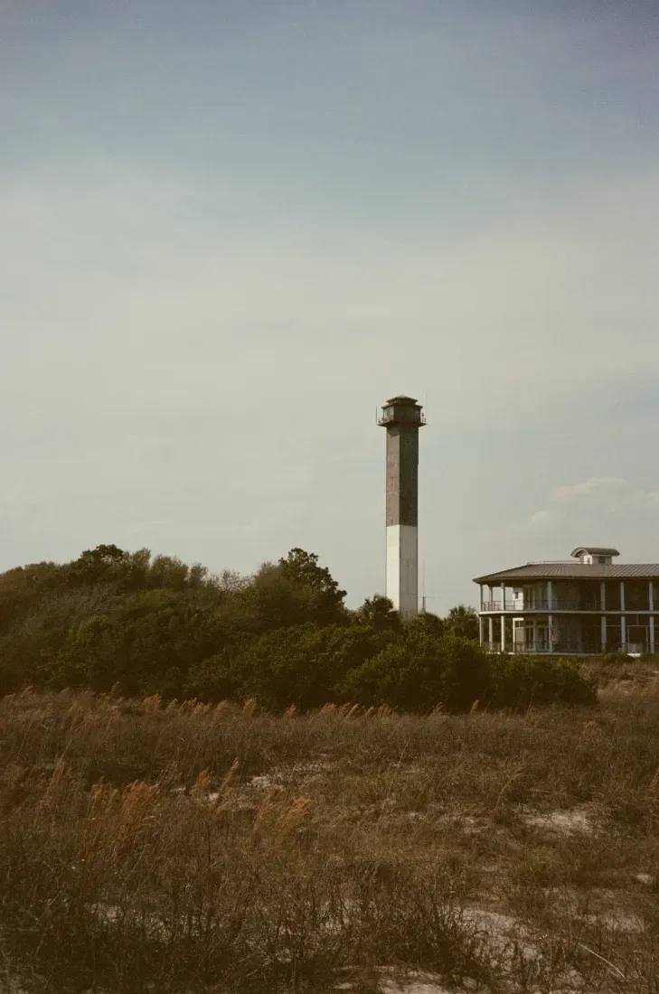Sulivan's Island Lighthouse rising above coastal shrubs with a low building under a cloudy sky