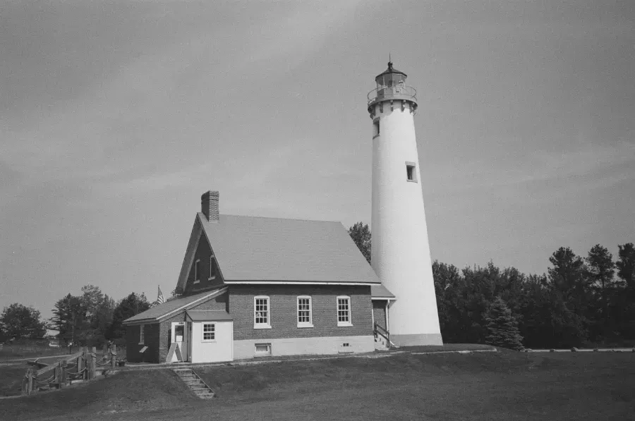 Tawas City Lighthouse with attached building on a grassy hill; trees and a blue sky in the background.
