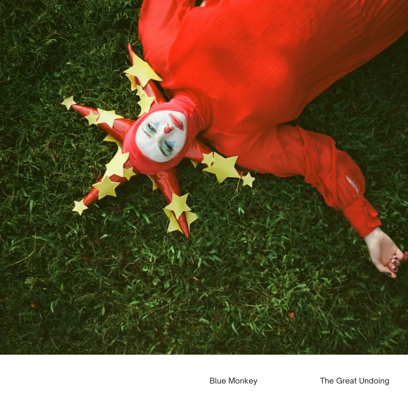 Charlee Remitz in red jester costume with yellow stars lies on green grass, looking upward. Album cover 